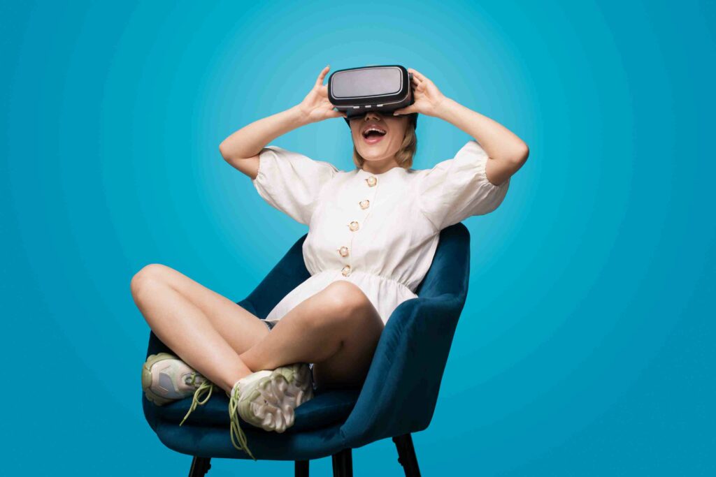 Blonde lady sitting in armchair is using virtual reality glasses and having fun on a blue studio wall